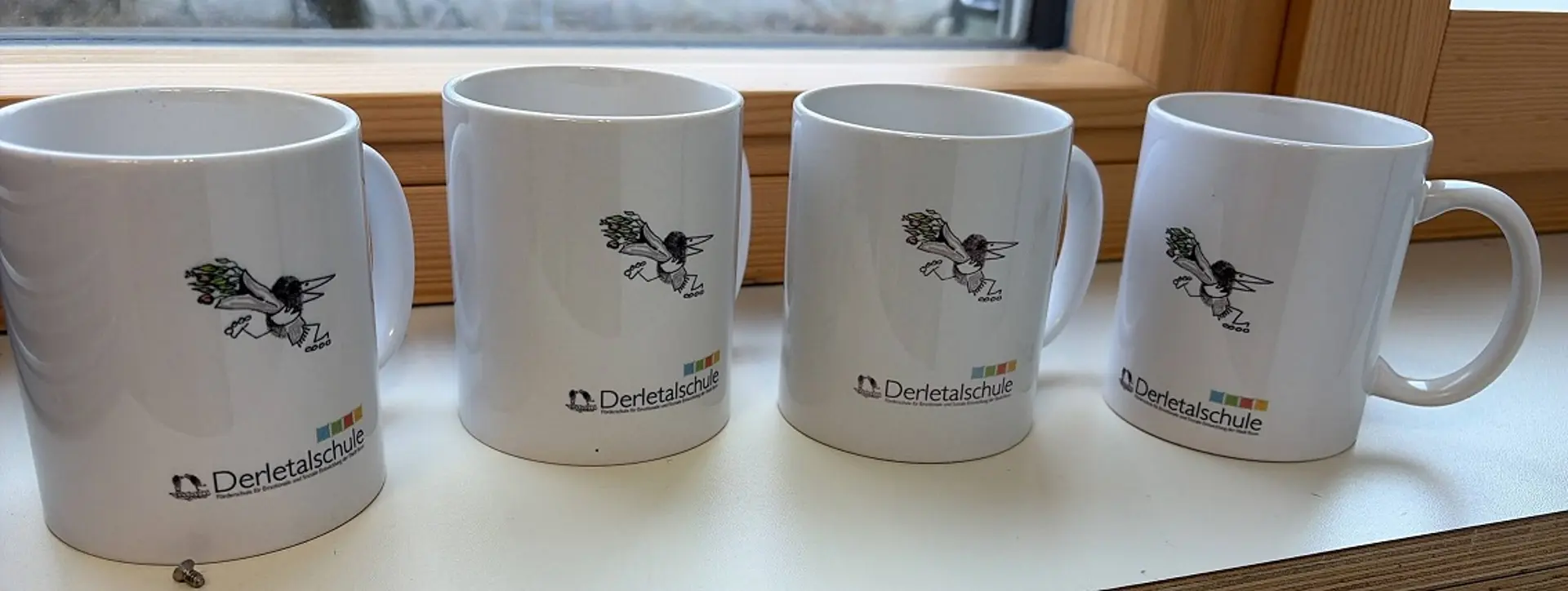 Vier Tassen, die auf einer Fensterbank nebeneinander gestellt sind. Auf jeder Tasse ist das Schrift-Logo der Derletalschule Bonn und eine Illustration von einem Rabe, der einen Blumenstrauß hält und Rollschuhe fährt, zu sehen. 