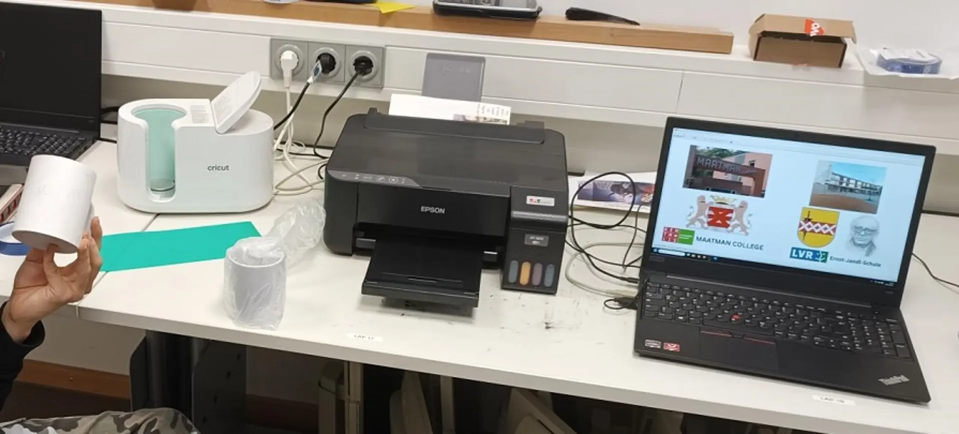 Ein Arbeitstisch in der Schule, darauf sind der Tassendrucker, ein Papierdrucker sowie ein aufgeklappter Laptop mit verschiedenen Logos und Bildern am leuchtenden Bildschirm zu sehen. 