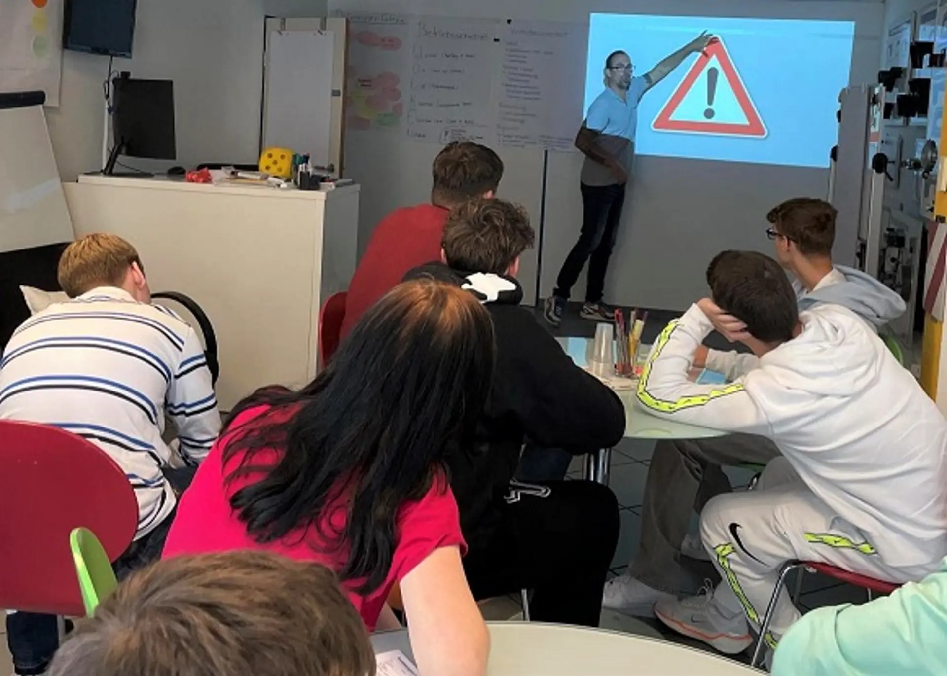 Eine Gruppe von Jugendlichen nimmt an einem Theorieunterricht in einer Fahrschule teil. Der Fahrlehrer zeigt an einer Projektionswand auf ein rotes Verkehrszeichen mit Ausrufezeichen für die Gefahrenstelle.