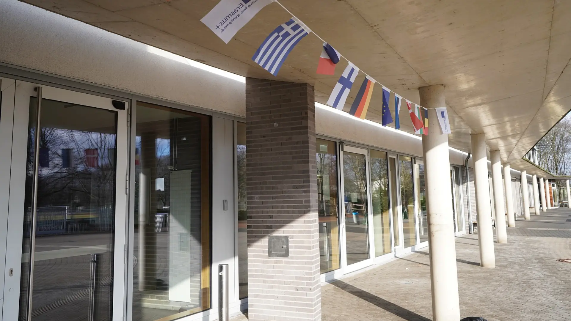 Der überdachte Eingangsbereich der Schule mit großen Glasfronten und hellen Säulen. Unter dem Vordach hängt eine bunte Wimpelkette mit verschiedenen europäischen Nationalflaggen und dem Erasmus+ Logo.