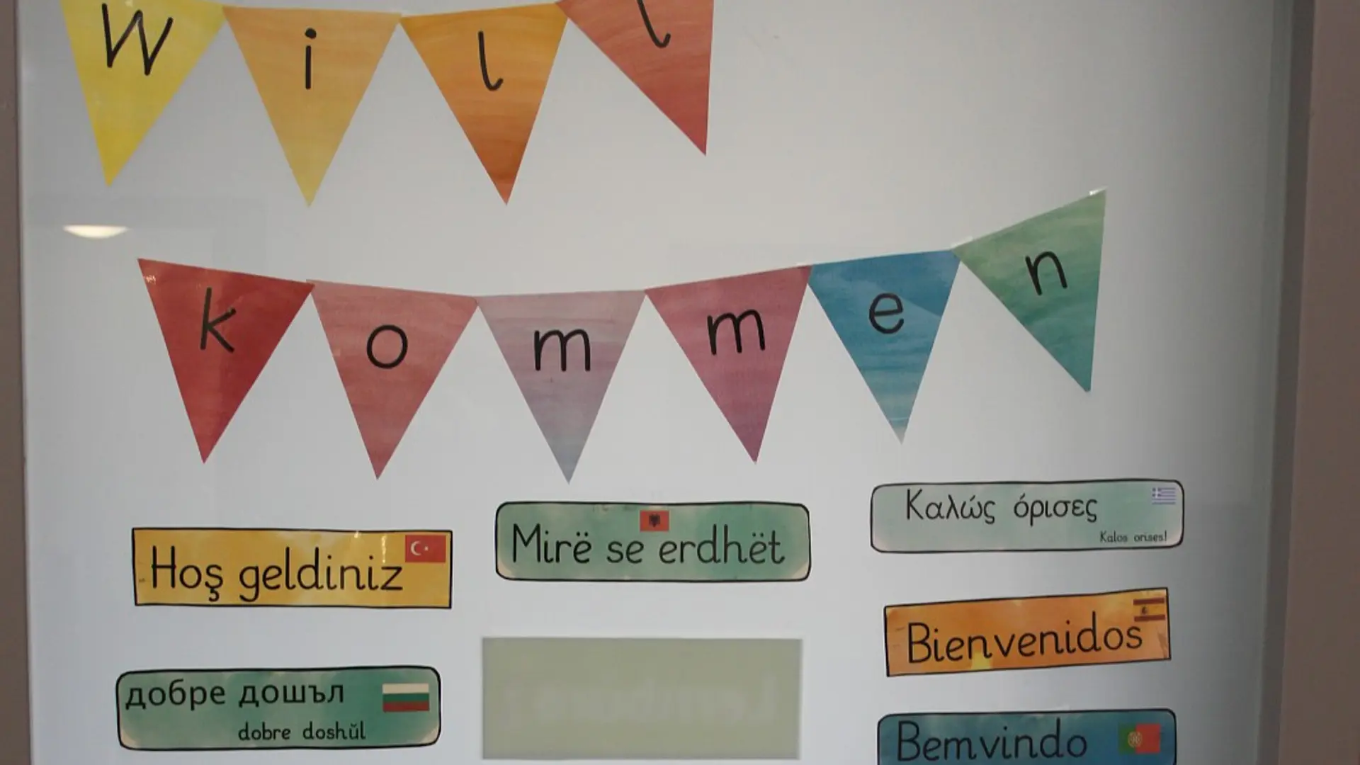 Eine Glastür in der Schule, die mit einer bunten Wimpelkette mit der Aufschrift "Willkommen" dekoriert ist. Darunter befinden sich zahlreiche Schilder, die das Wort "Willkommen" in vielen verschiedenen Sprachen.
