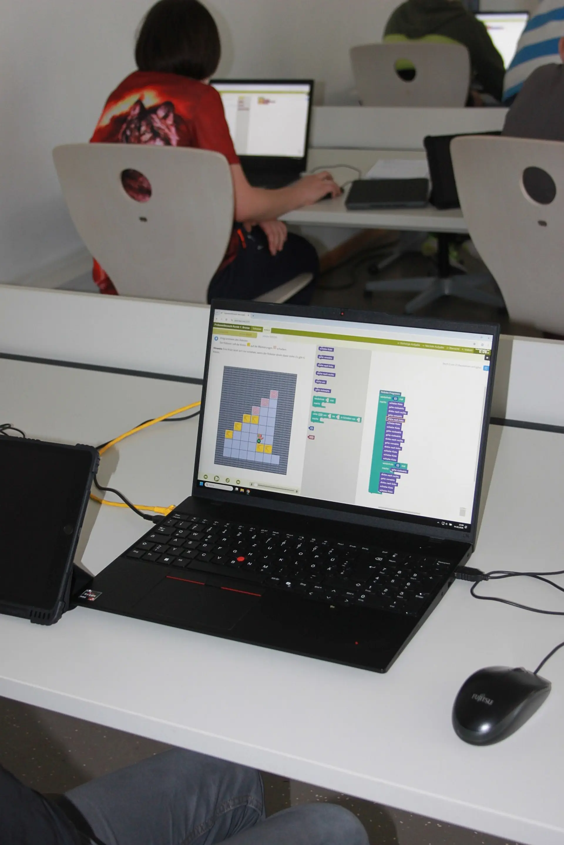 Nahaufnahme eines Laptops auf einem Schreibtisch in einem Klassenzimmer. Auf dem Bildschirm ist eine Lernsoftware für visuelle Programmierung zu sehen. Im Hintergrund sitzen weitere Schüler*innen konzentriert an ihren Laptops und Tablets.